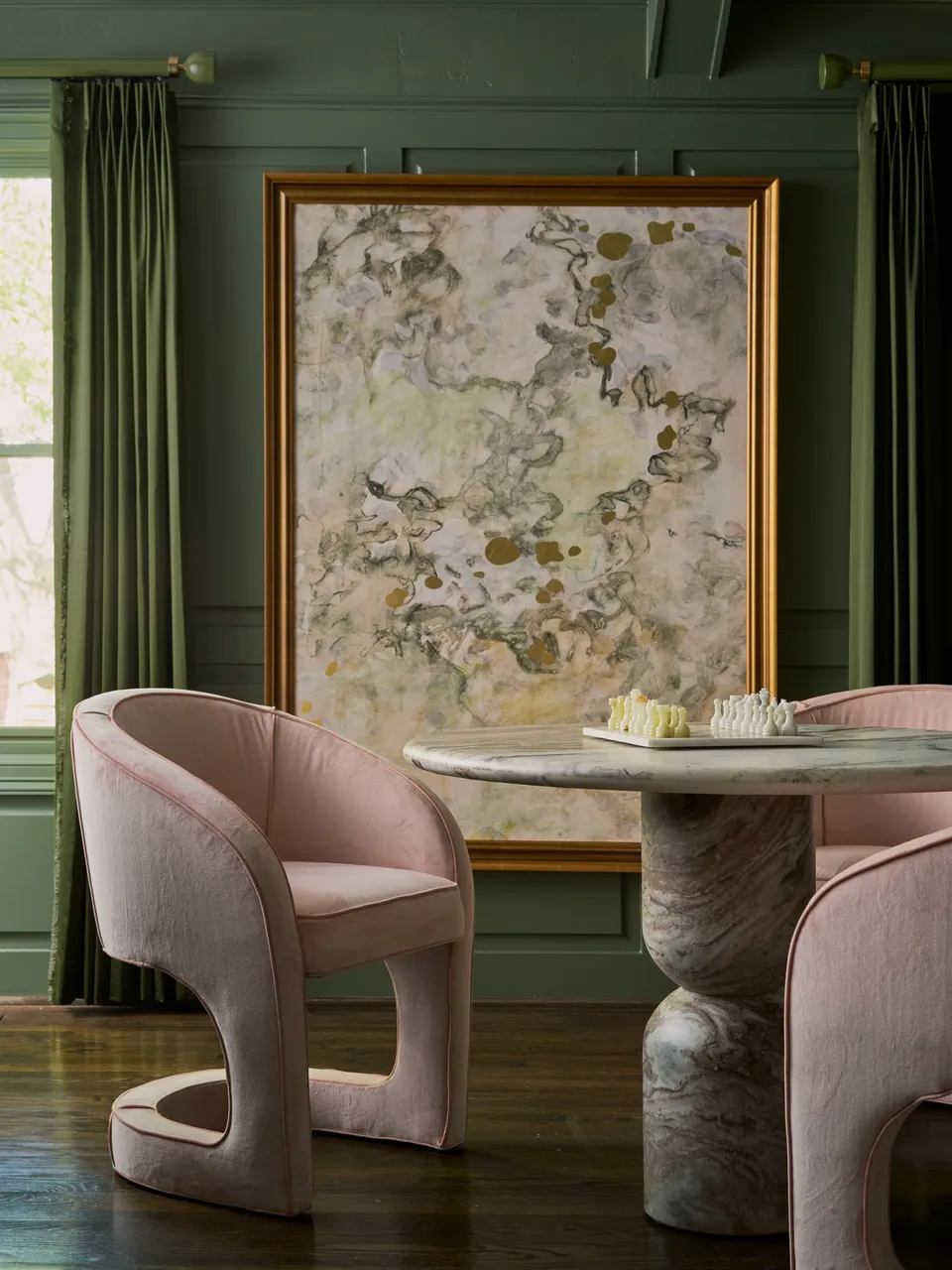 Dining corner with green panelled walls, a large framed abstract painting, blush upholstered chairs and a round marble pedestal table