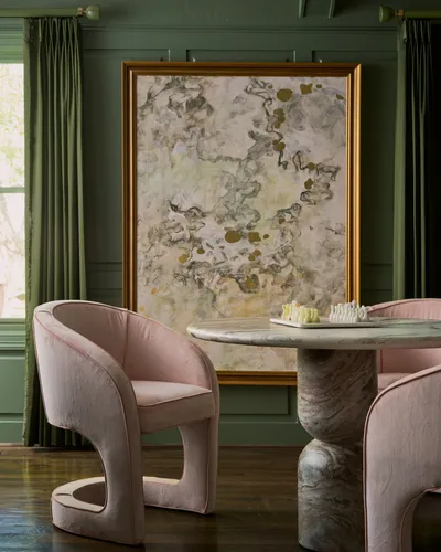 Dining nook with green panelled walls, a large framed abstract painting and blush upholstered chairs around a marble pedestal table.