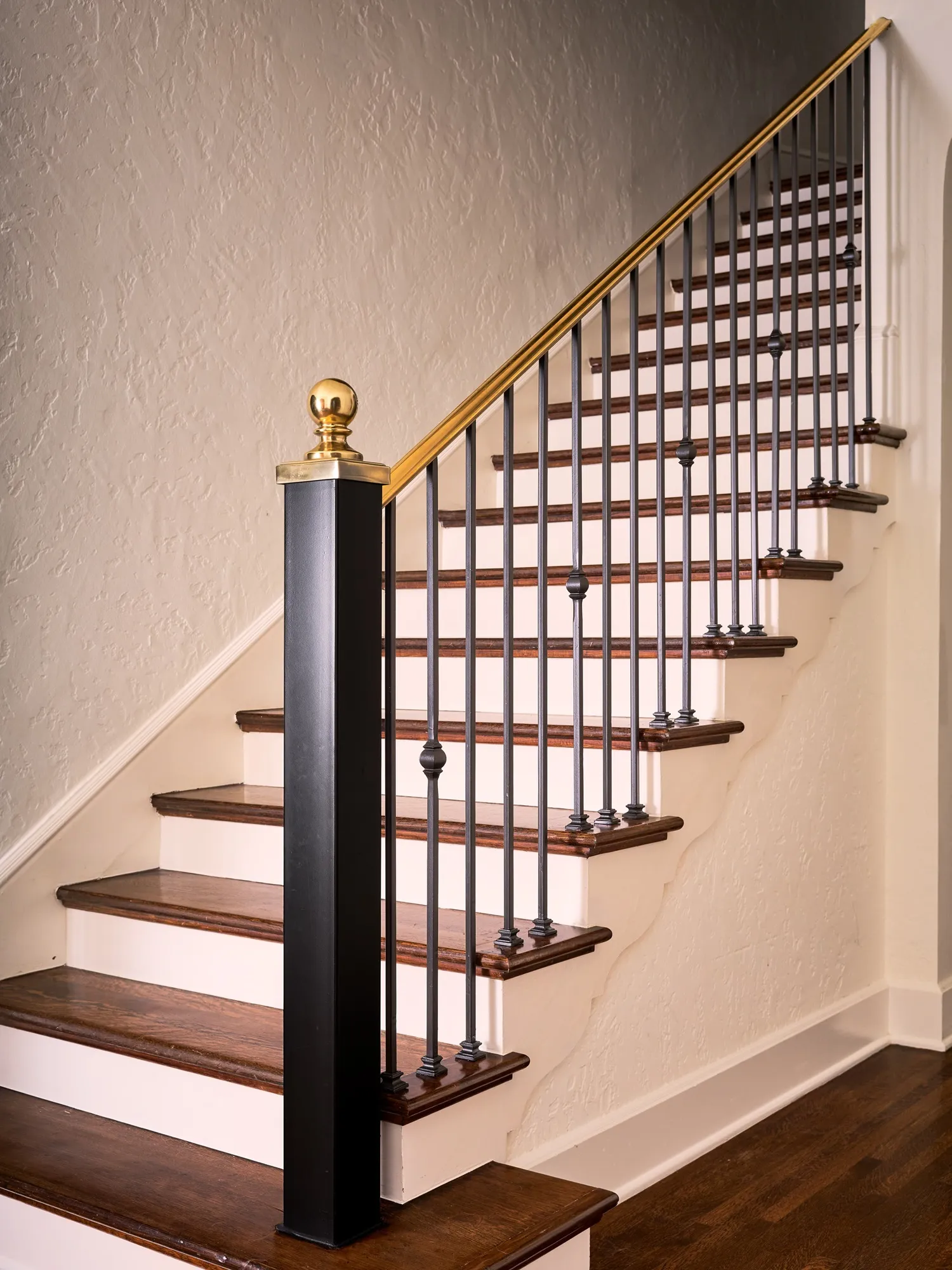 Staircase with dark wood treads, wrought iron balusters, a brass handrail and a black newel post against a textured white wall