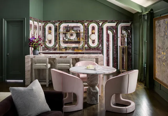 Green lounge and bar area with a patterned marble backsplash, brass shelving and pink velvet chairs around a round marble table