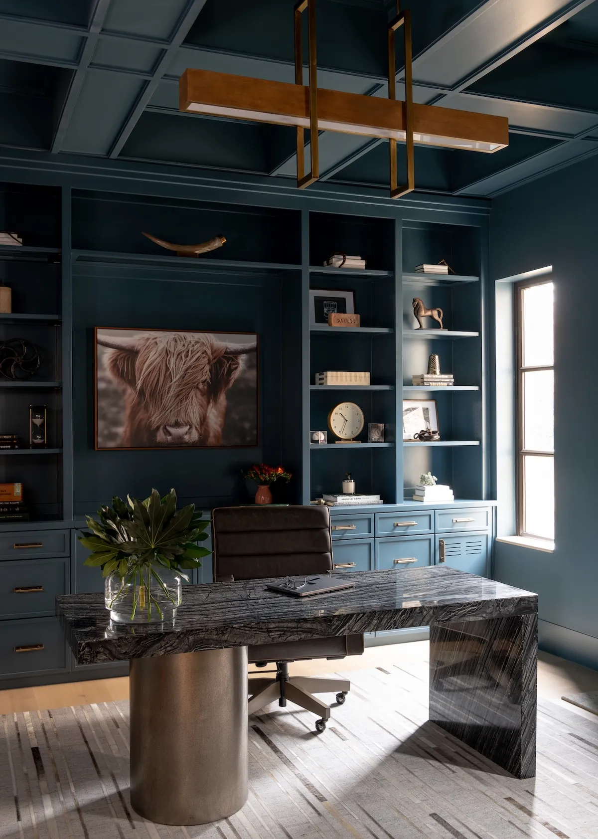 home office with deep blue built-in shelving, a highland cow portrait, a brass pendant light and a black marble desk.