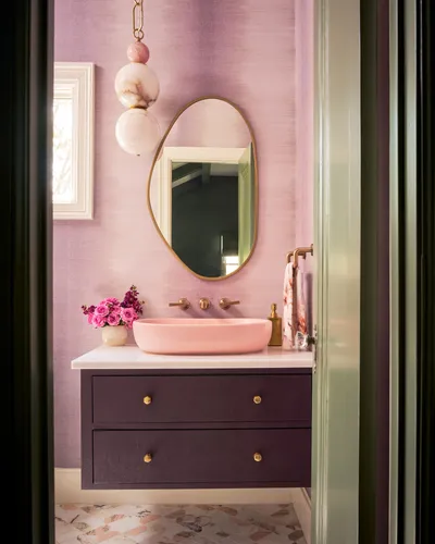 Powder room with lilac grasscloth walls, an organic brass mirror, a pink vessel sink and a plum vanity with brass knobs.