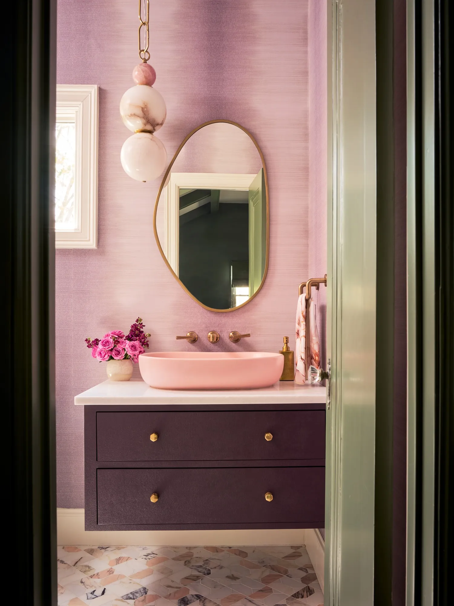 Bathroom with lilac walls, an organic gold-framed mirror, a pink basin on a plum vanity, a pearl pendant light and pink roses