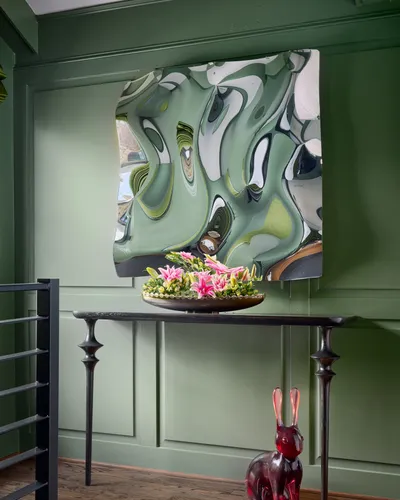 Sage green panelled entry with a sculptural mirrored wall piece above a black console table with pink lilies and a red glass rabbit.