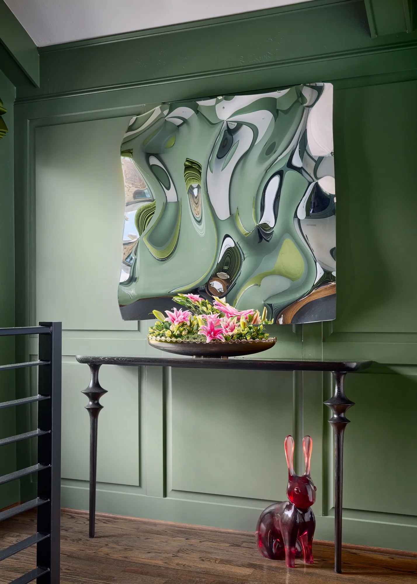 Landing with green panelled walls, a mirrored abstract artwork above a slim black console with pink flowers, and a red glass rabbit sculpture on the floor
