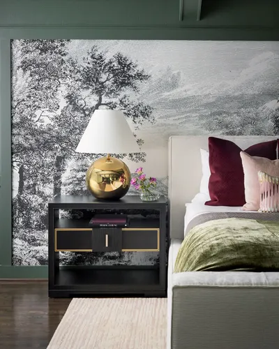 Bedroom corner with a black and white landscape mural, a gold globe lamp on a black nightstand and an upholstered bed with burgundy and blush pillows.