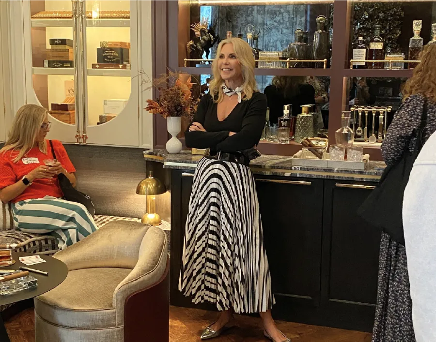 Traci Connell smiling with crossed arms beside a bar cabinet lined with glass decanters.