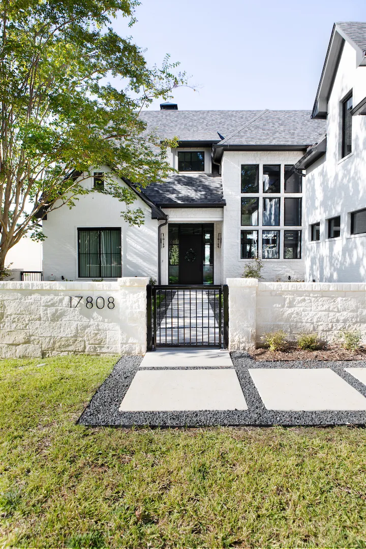 white brick house with black windows behind a limestone wall, an iron gate and concrete pavers across the lawn.