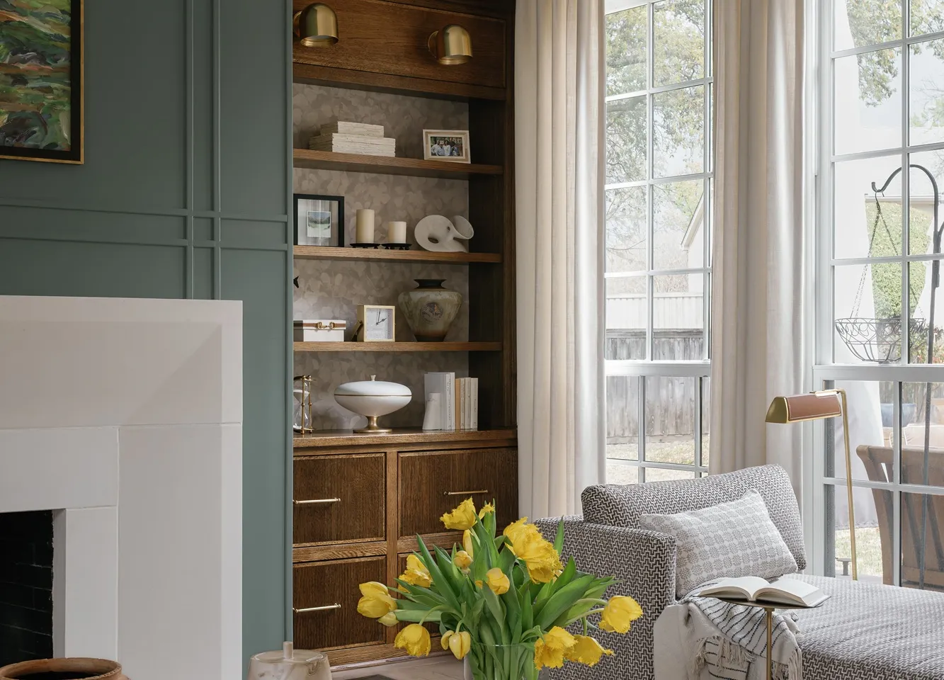Living room with sage green panelled walls, wooden built-in shelving, a limestone fireplace and yellow tulips on a dark coffee table