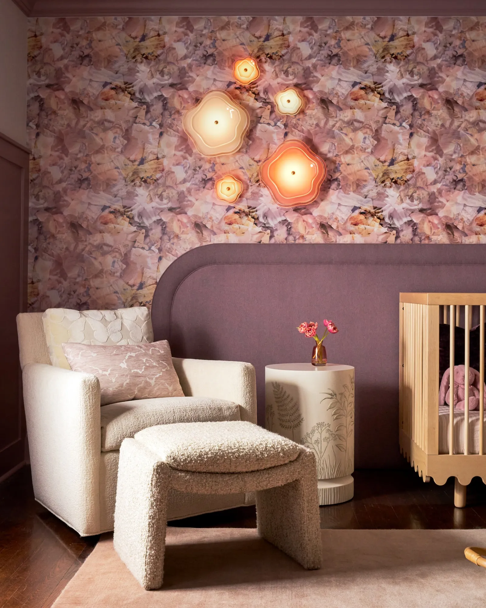 Nursery corner with pink floral wallpaper, sculptural glowing wall lights, a cream armchair with an ottoman, a small side table and a wooden crib