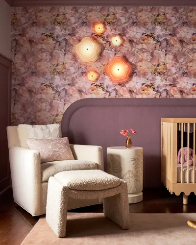 Nursery corner with floral wallpaper and flower-shaped wall lights, a cream bouclé armchair and ottoman, a botanical side table and a wooden crib.