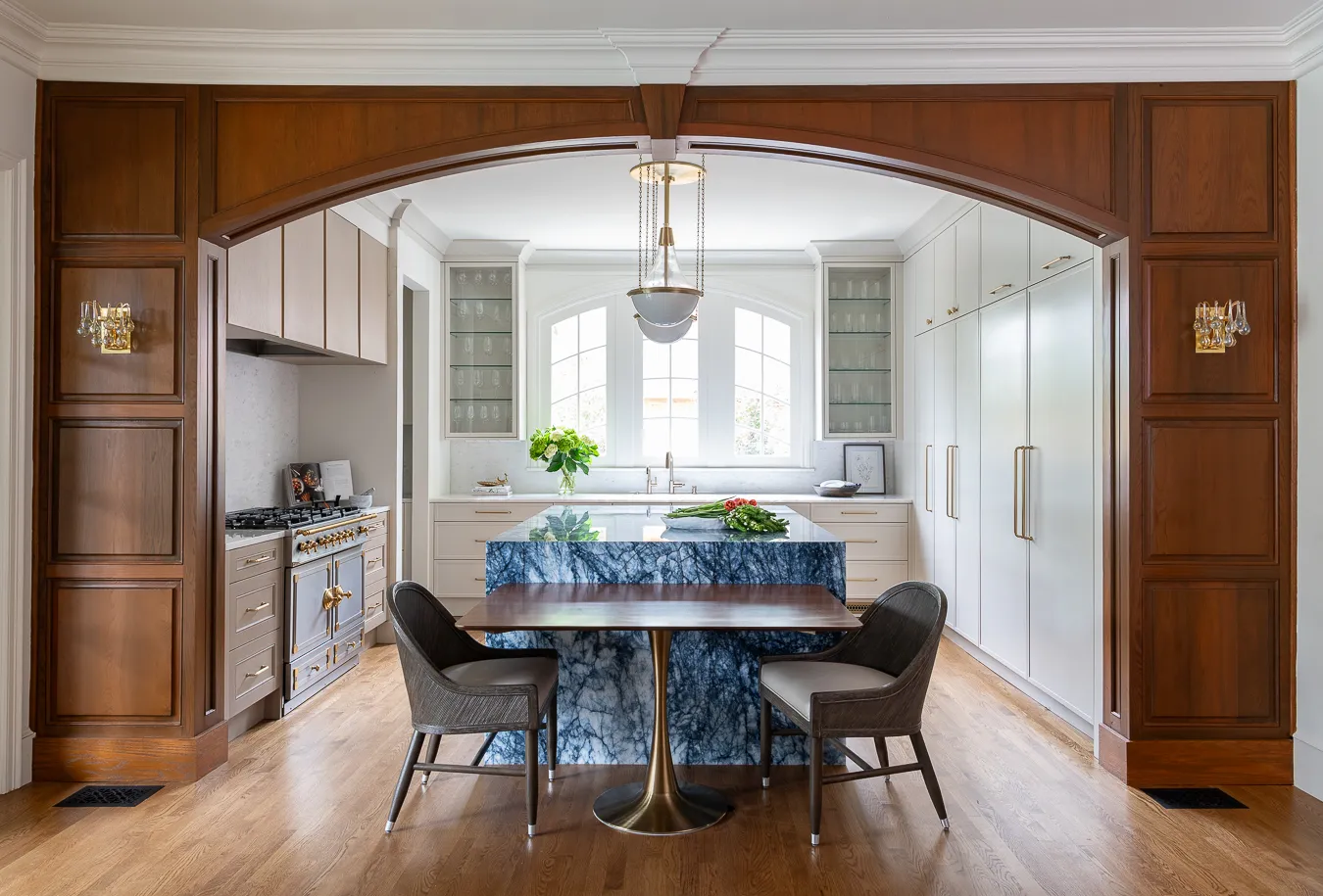 Kitchen seen through a wooden arch, with a blue marble island, attached walnut dining table and a pendant light over white cabinetry
