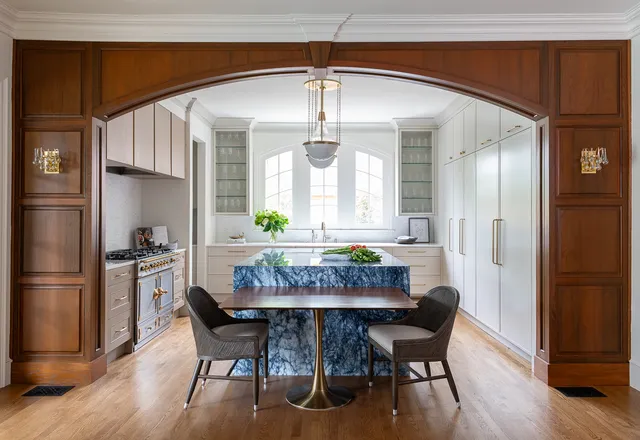 Kitchen seen through a wooden arch, with a blue marble island, attached walnut dining table and a pendant light over white cabinetry