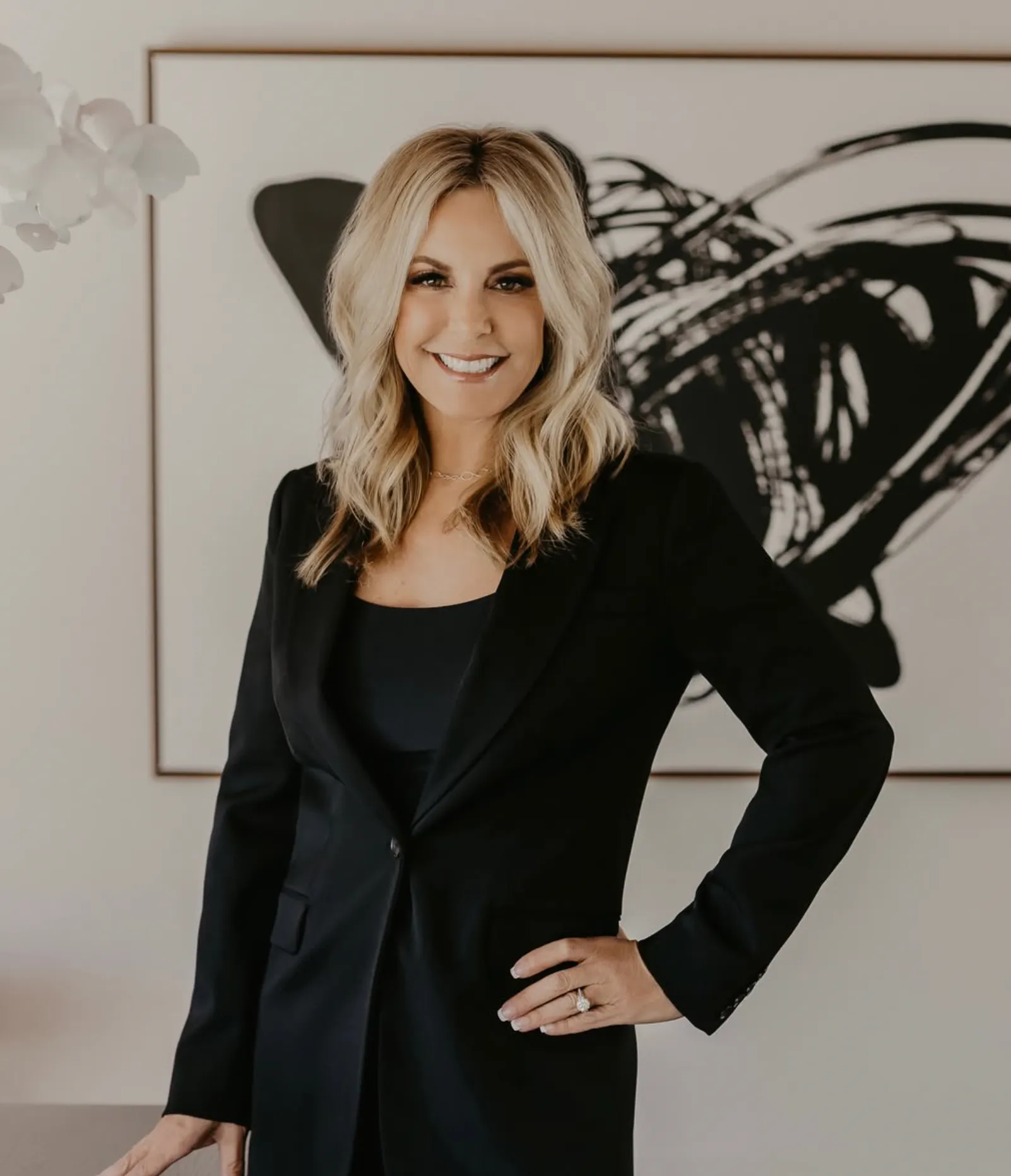 Smiling blonde woman in a white top and navy floral pencil skirt posing beside a dark console table with decorative accessories.