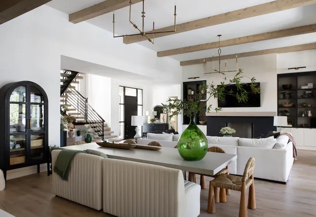 Open plan living and dining space with exposed wooden ceiling beams, brass chandeliers, white sofas and a black fireplace wall