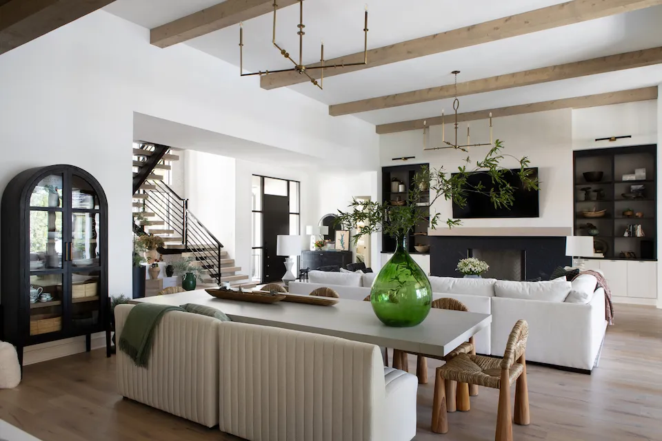 Open plan living and dining space with exposed wooden ceiling beams, brass chandeliers, white sofas and a black fireplace wall
