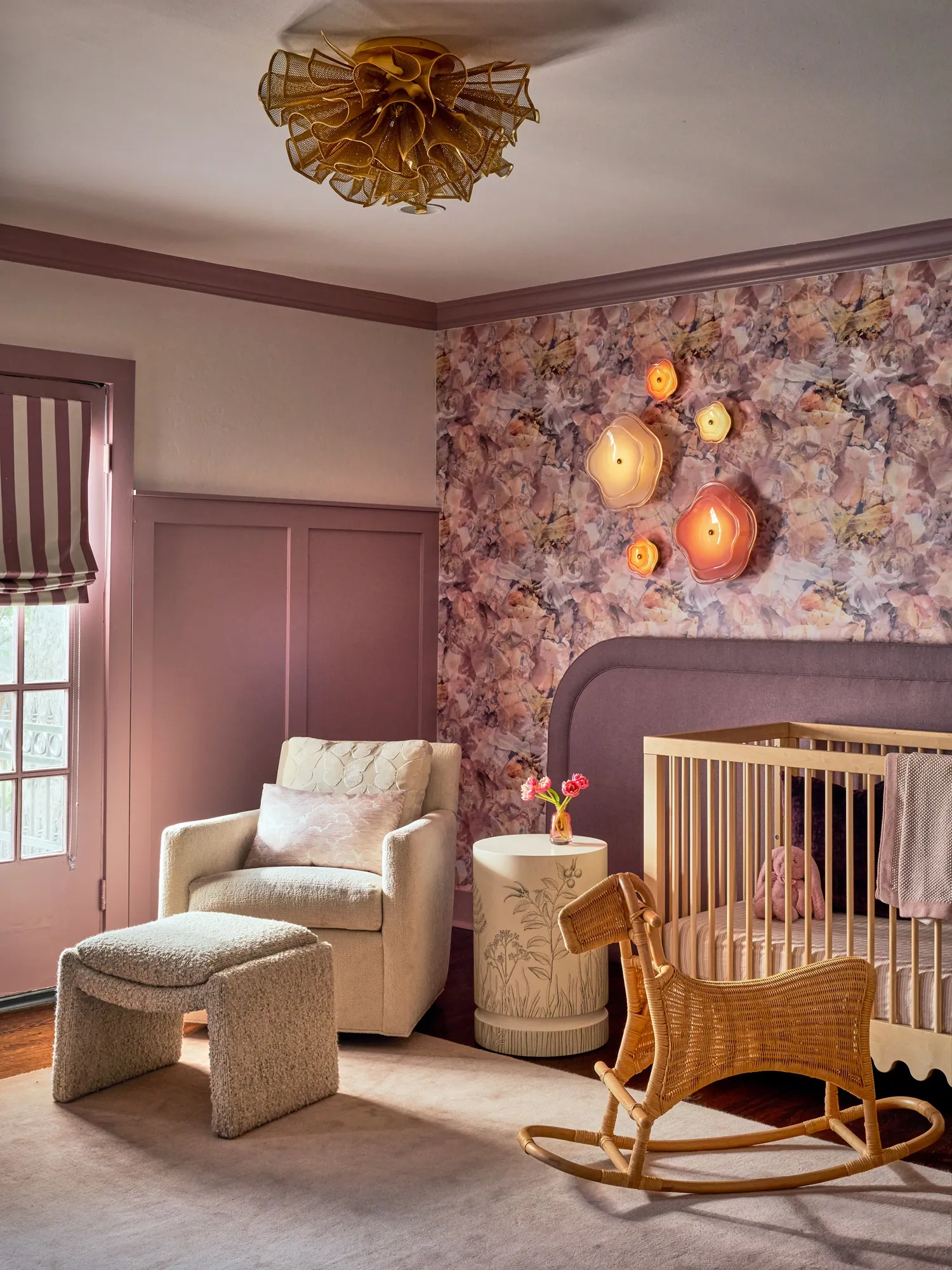 Nursery with mauve panelled walls and floral wallpaper, a gold flower-shaped ceiling light, a cream armchair, a wooden crib and a woven rocking horse