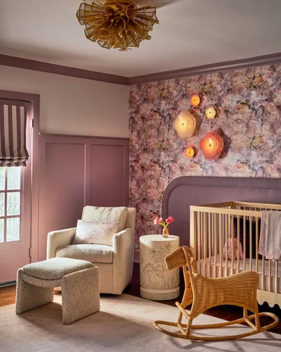 Mauve nursery with a ruffled gold ceiling light, floral wallpaper, a cream armchair, a wooden crib and a rattan rocking horse.