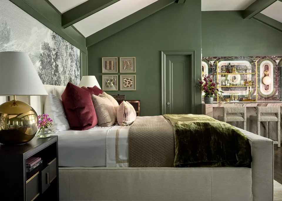 Bedroom under a sloped ceiling with green beams, a scenic mural wall, burgundy and blush cushions, a green velvet throw and a brass table lamp