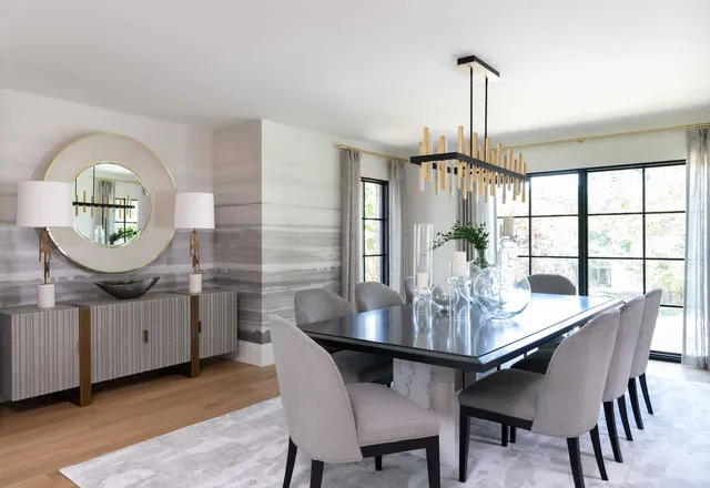 Dining room with a black table and grey upholstered chairs, a linear brass chandelier, round mirror over a sideboard and hand painted walls