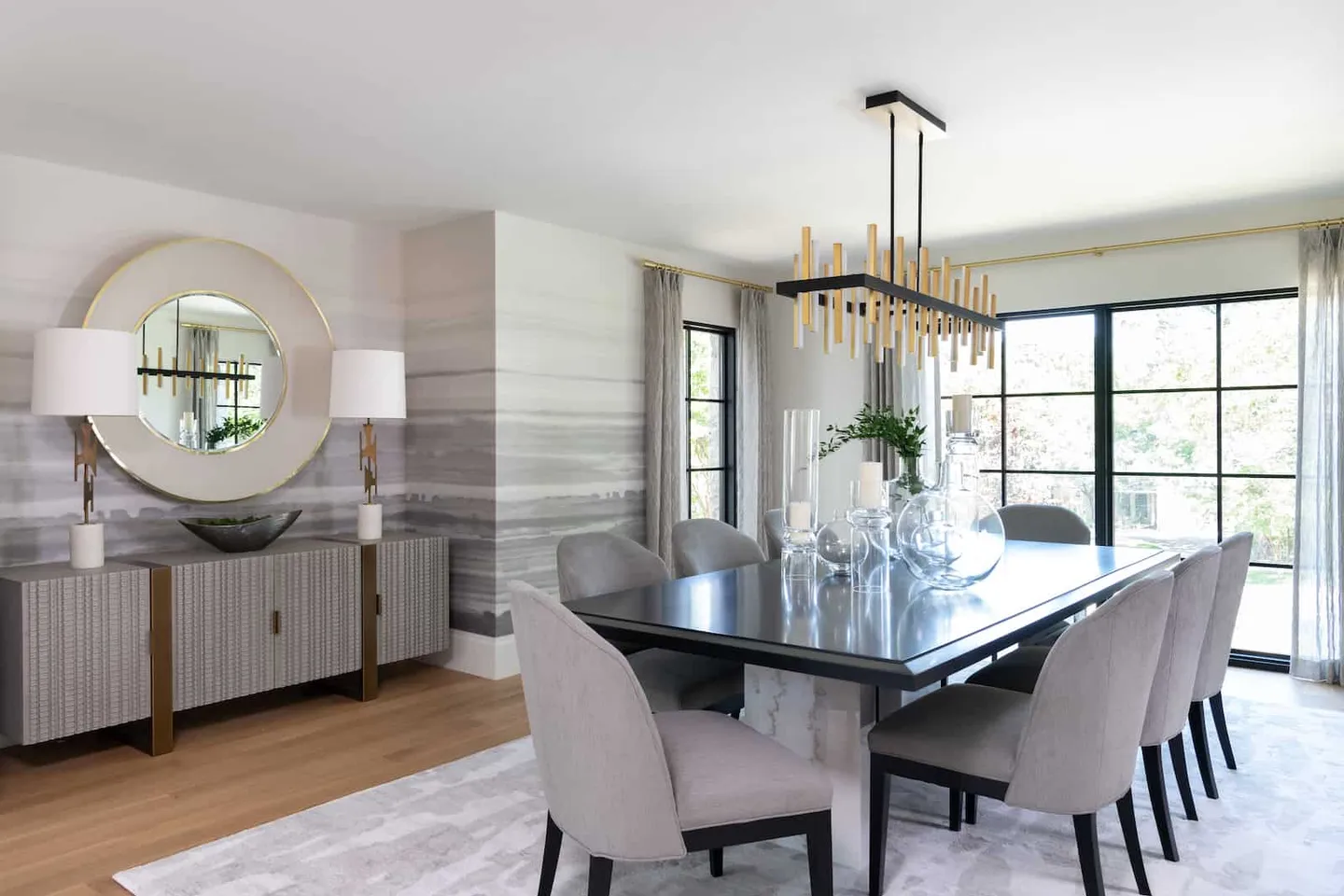 Dining room with a black table and grey upholstered chairs, a linear brass chandelier, round mirror over a sideboard and hand painted walls