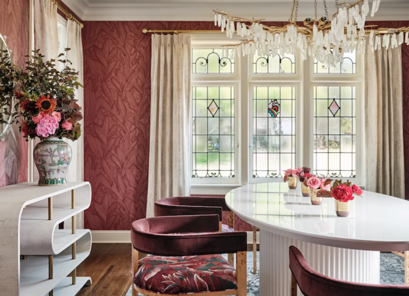 Dining room with red patterned wallpaper, leaded glass windows, a quartz chandelier and burgundy velvet chairs around an oval white table