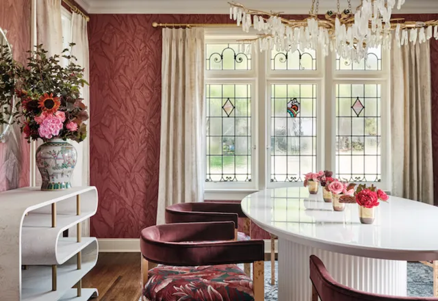 Dining room with red patterned wallpaper, leaded glass windows, a quartz chandelier and burgundy velvet chairs around an oval white table